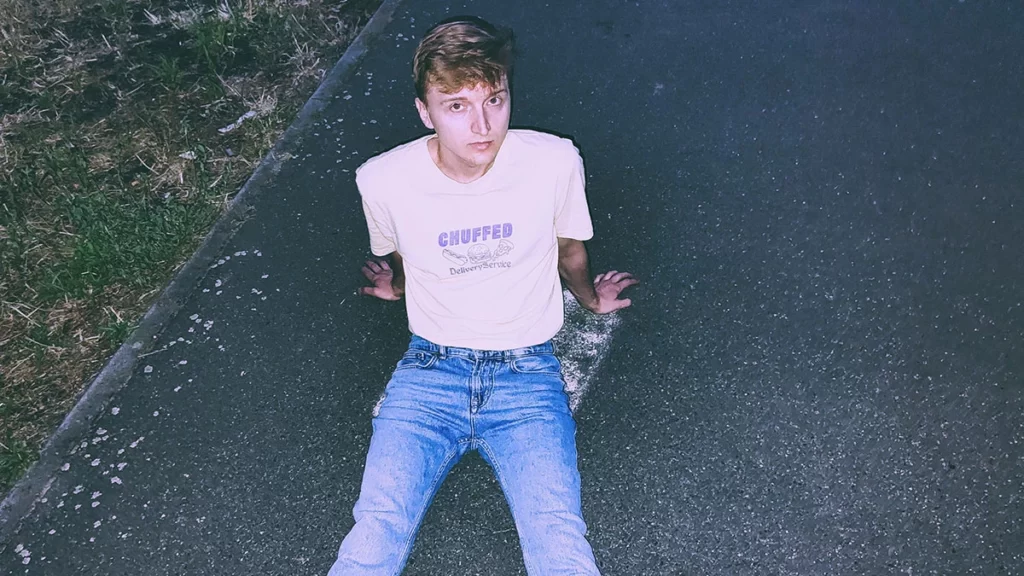 Picture of Patryk Kania sitting on the asphalt , legs stretched out, he has jeans and light pink t-shirt. The t-shirt has this written on top Chuffed - below icons of hotdog, hamburger and pizza slice - below the icons it says Delivery service. Patryk Kania has dark brown hair and brown eyes.