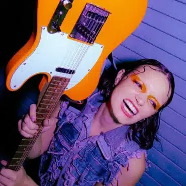 Ella Galvin holding a guitar upside down, against a purple background, wearing a jeans jacket.