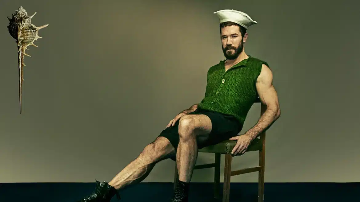 Zach Blew sits in a kitchen chair, wearing shorts, a green button-up shirt, a sailor hat, and black shoes. He has dark brown hair and a beard. Photo by Jon Jacobsen