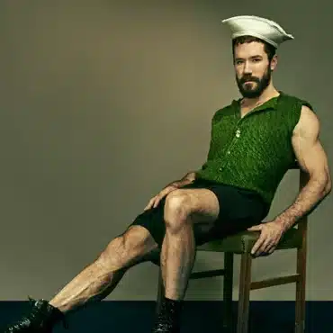 Zach Blew sits in a kitchen chair, wearing shorts, a green button-up shirt, a sailor hat, and black shoes. He has dark brown hair and a beard. Photo by Jon Jacobsen